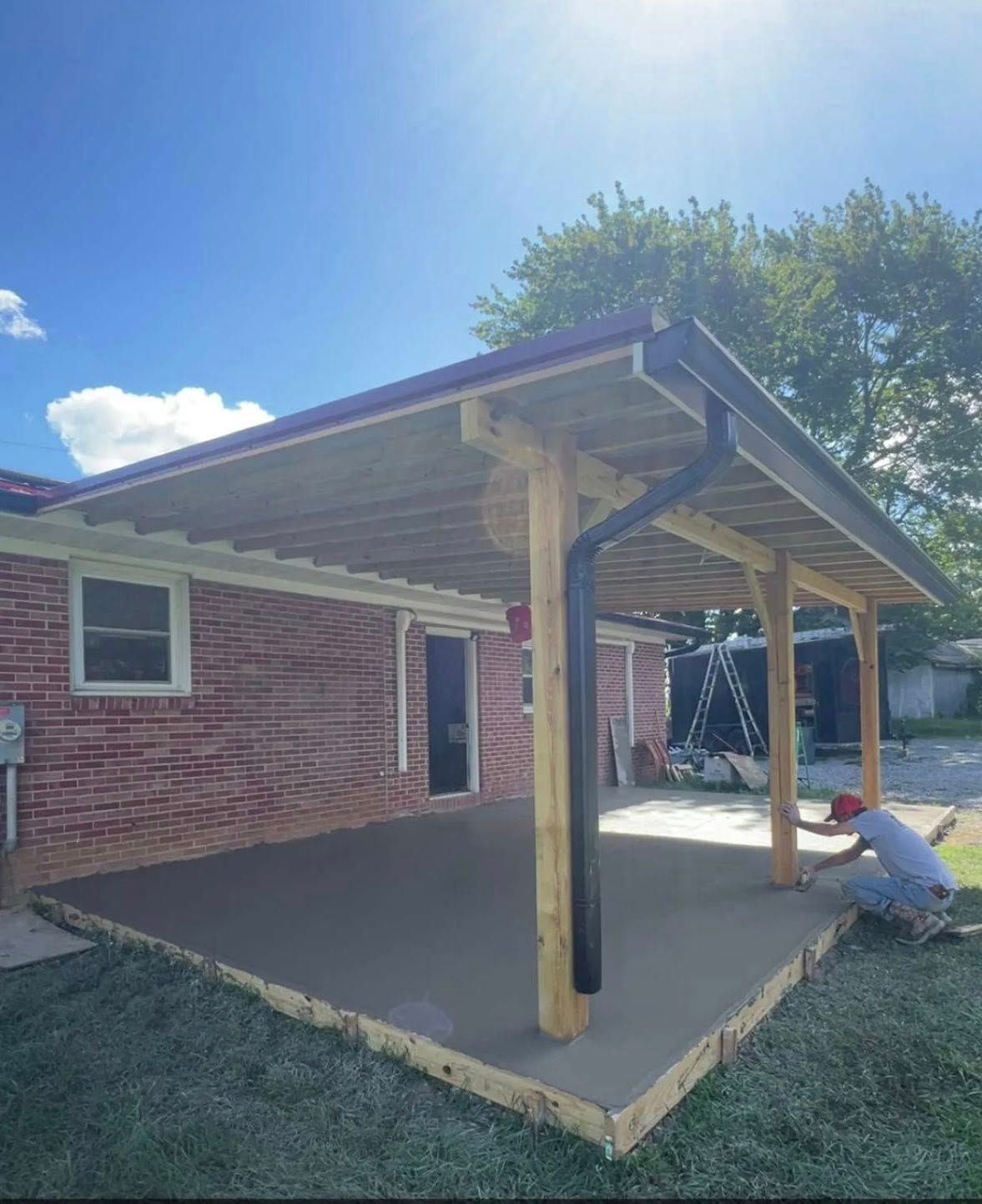 A person kneeling on a newly poured concrete patio beneath a wooden porch roof attached to a brick house.