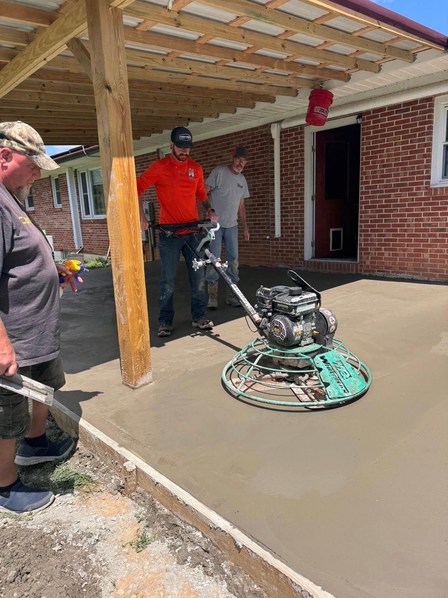 Three workers operate a power trowel to smooth a freshly poured concrete patio under a wooden covered porch.