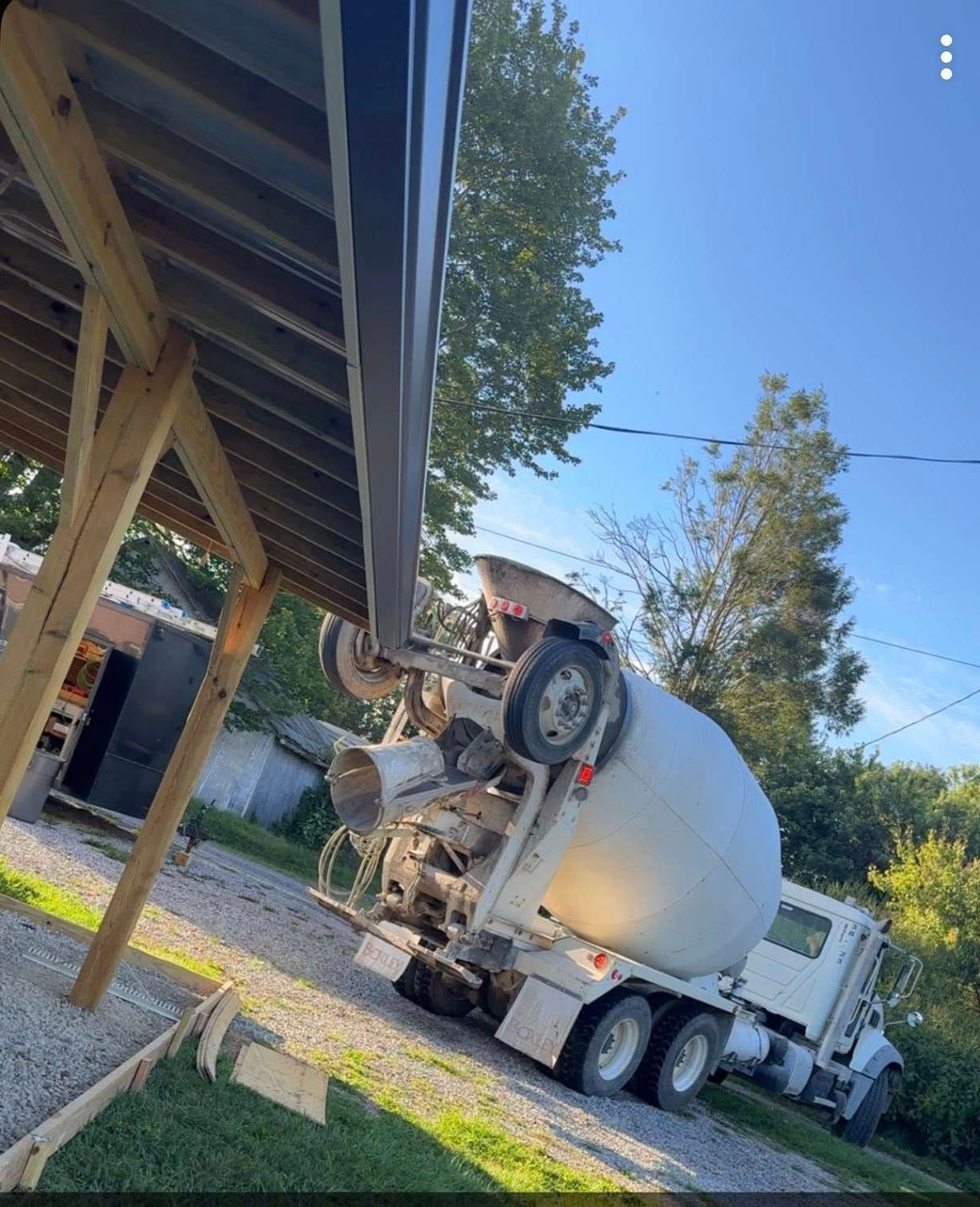 A concrete mixer truck parked on a gravel driveway beneath the edge of a wooden structure roof.