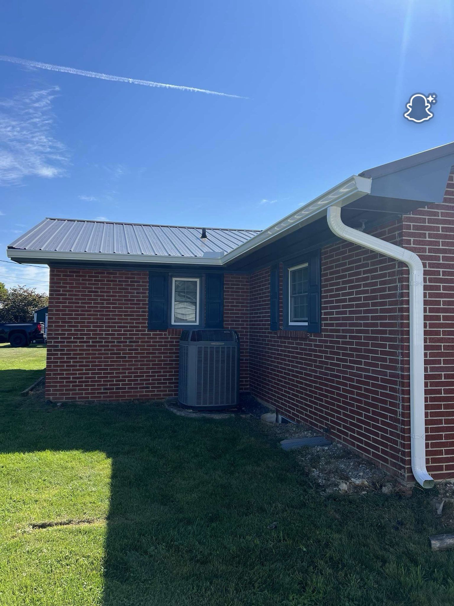A red brick house with a white gutter downspout, metal roof, and an outdoor air conditioning unit on a grassy lawn.
