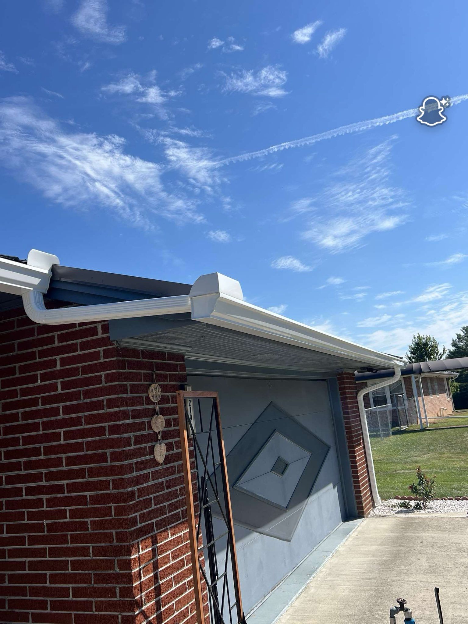 White gutters installed on the exterior corner of a brick house under a blue, sunny sky with a contrail.