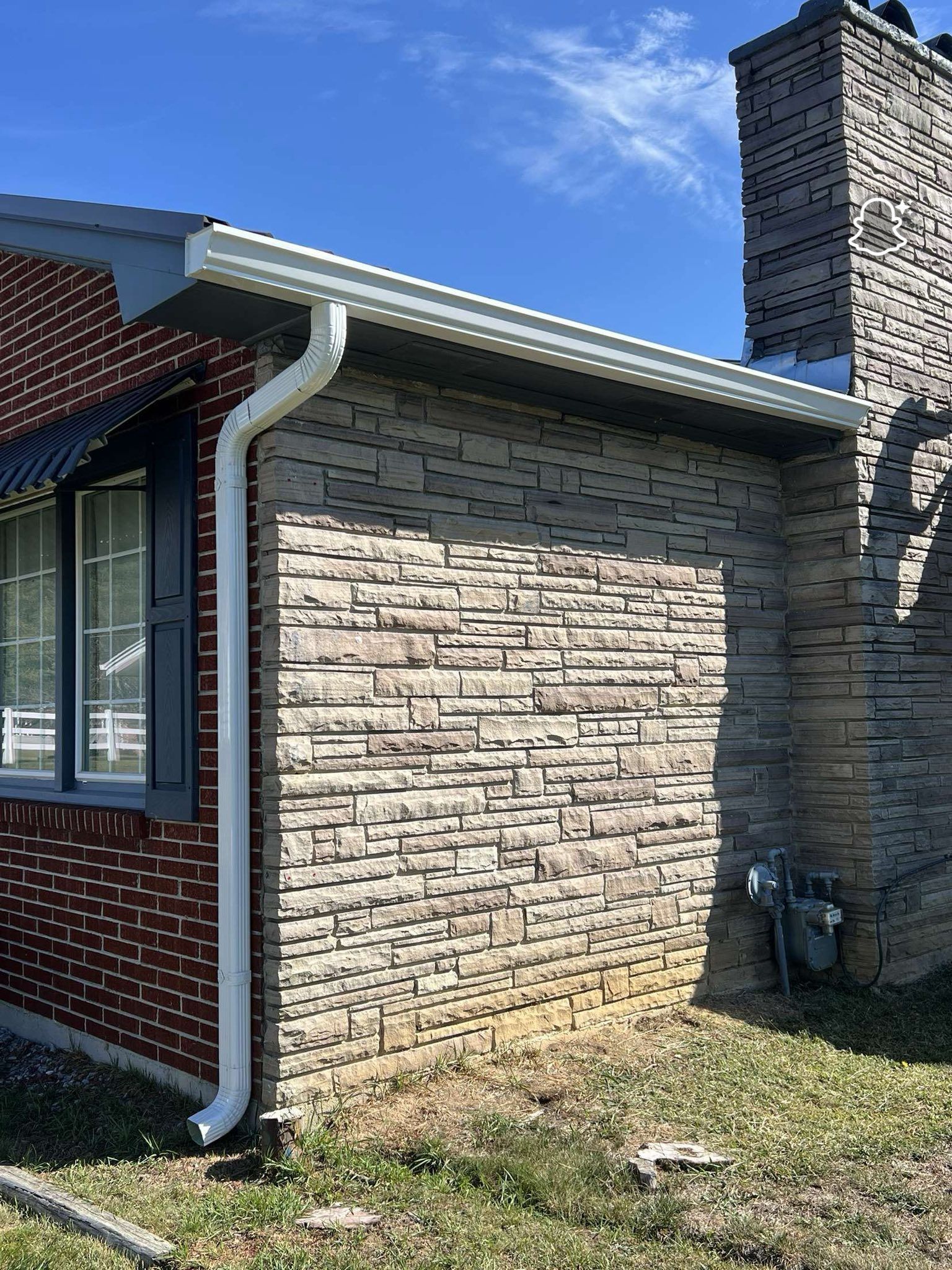 A side view of a brick and stone house exterior with a chimney, white gutter, and a downspout against a blue sky.
