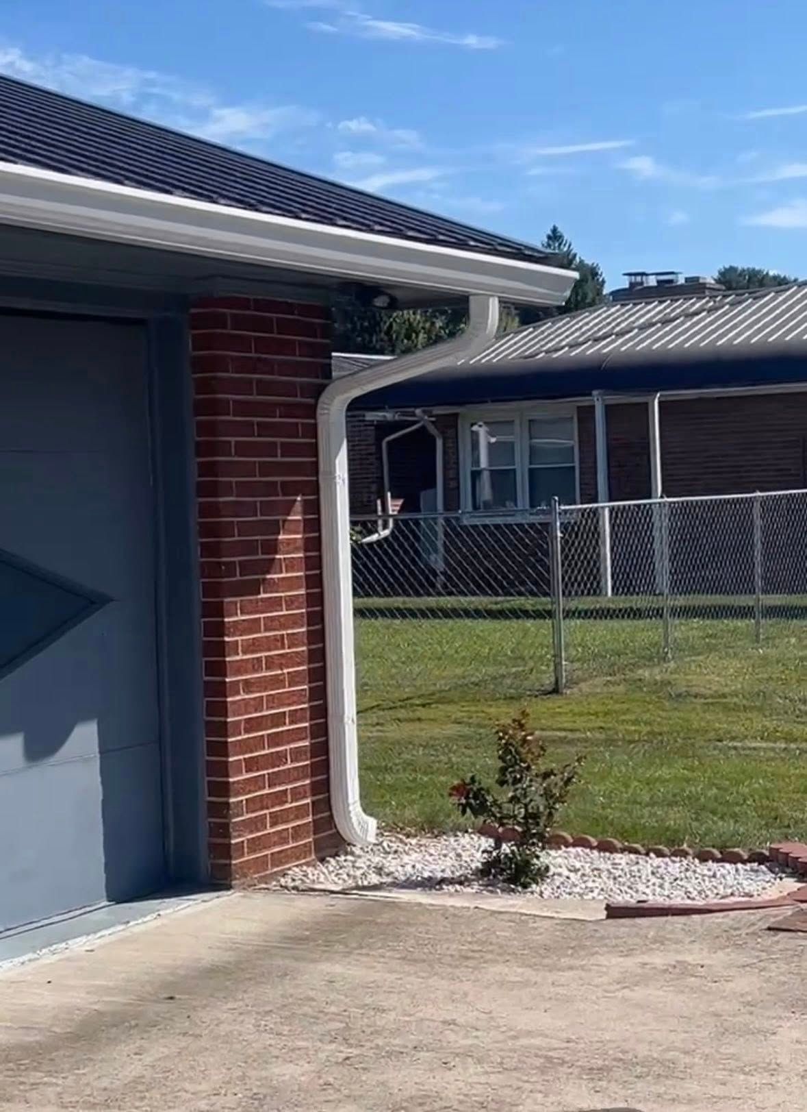 A white downspout on a brick corner of a house next to a small plant in a rock bed, with a neighboring house in the back.