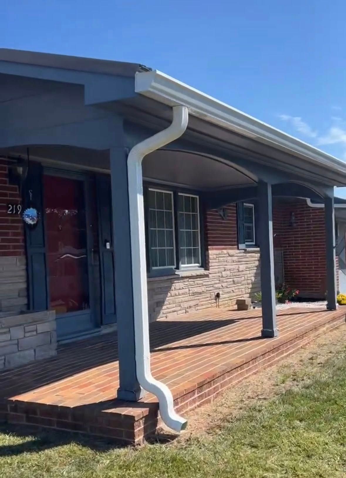 A front porch with a red door, brick floor, stone accent wall, grey support posts, and a white downspout on a sunny day.