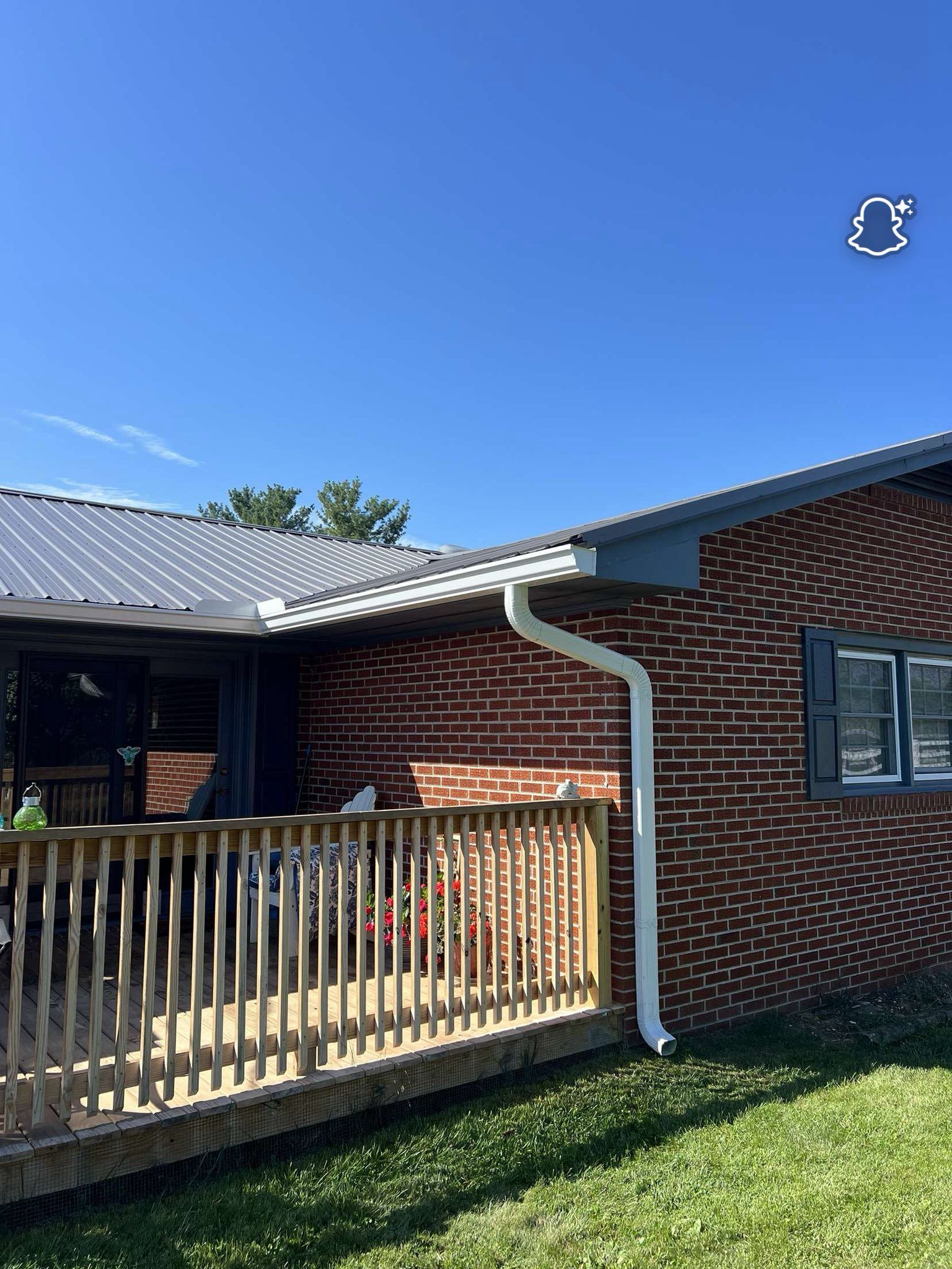 A red brick house exterior features a wooden porch, a white gutter downspout, and a dark metal roof under a blue sky.