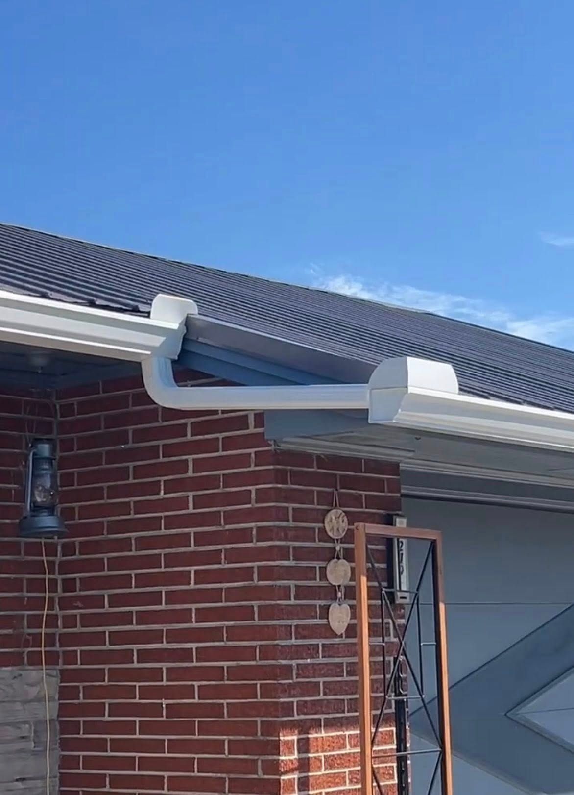 A white metal gutter system attached to the roofline of a red brick house under a clear blue sky.