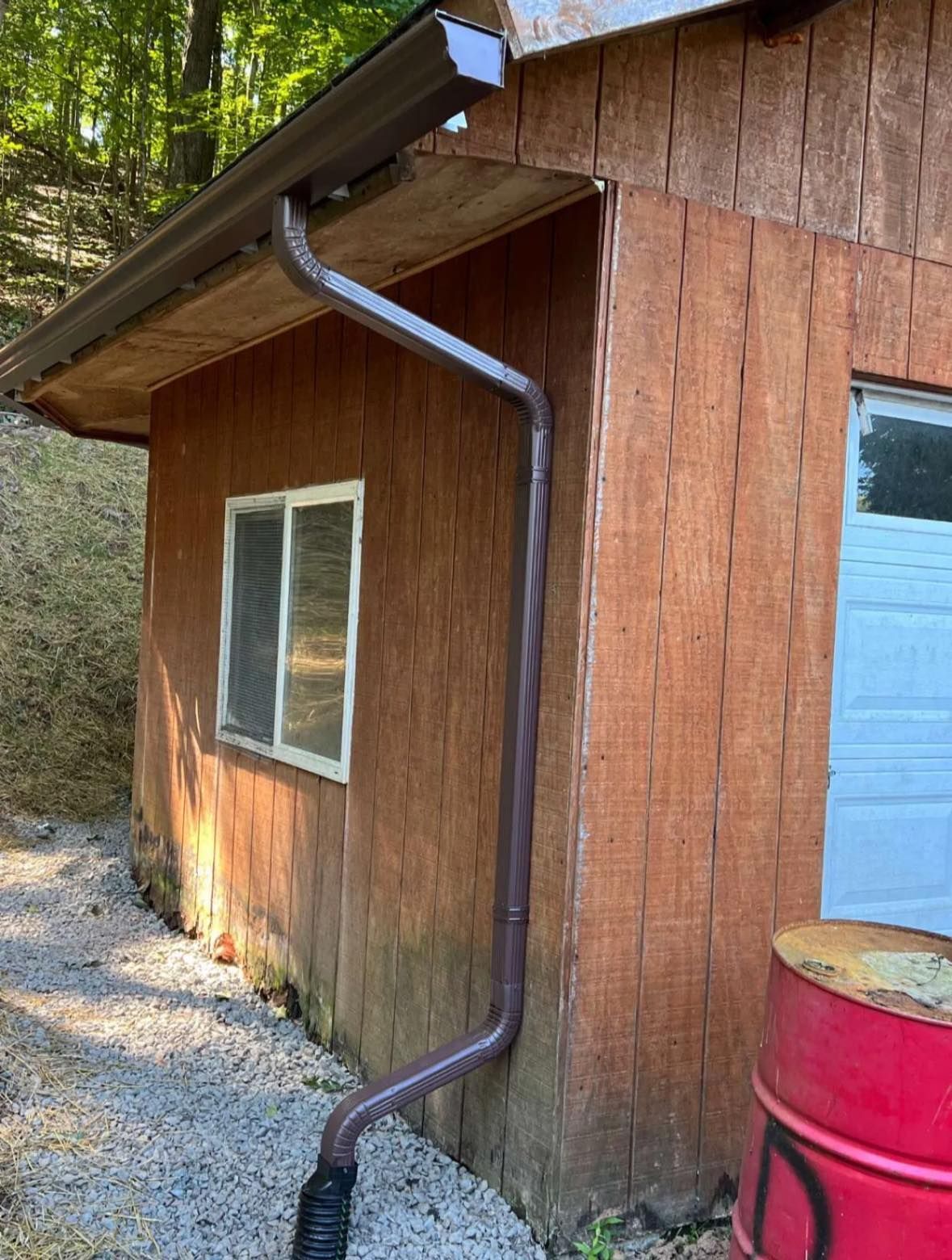 A brown gutter downspout attached to a wooden building, extending down to a black corrugated drain pipe on gravel ground.