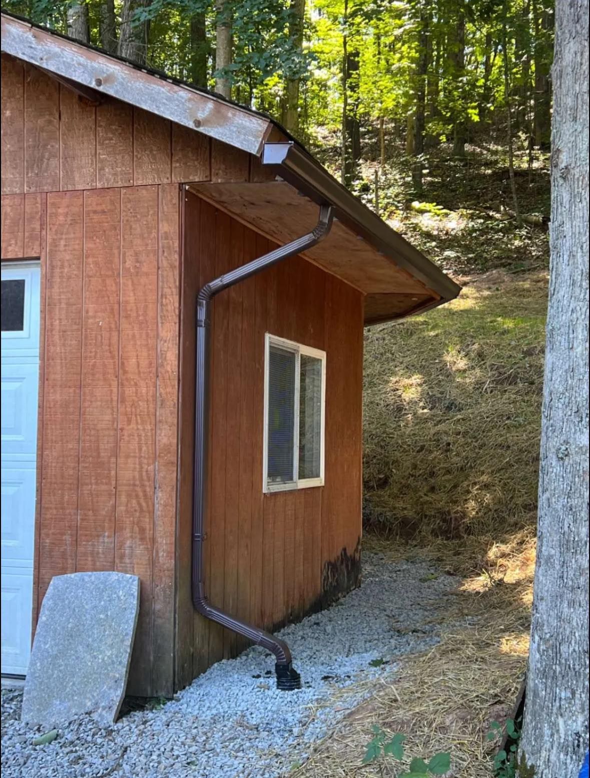 A brown wooden shed corner featuring a dark brown gutter downspout system discharging onto a gravel ground surface.