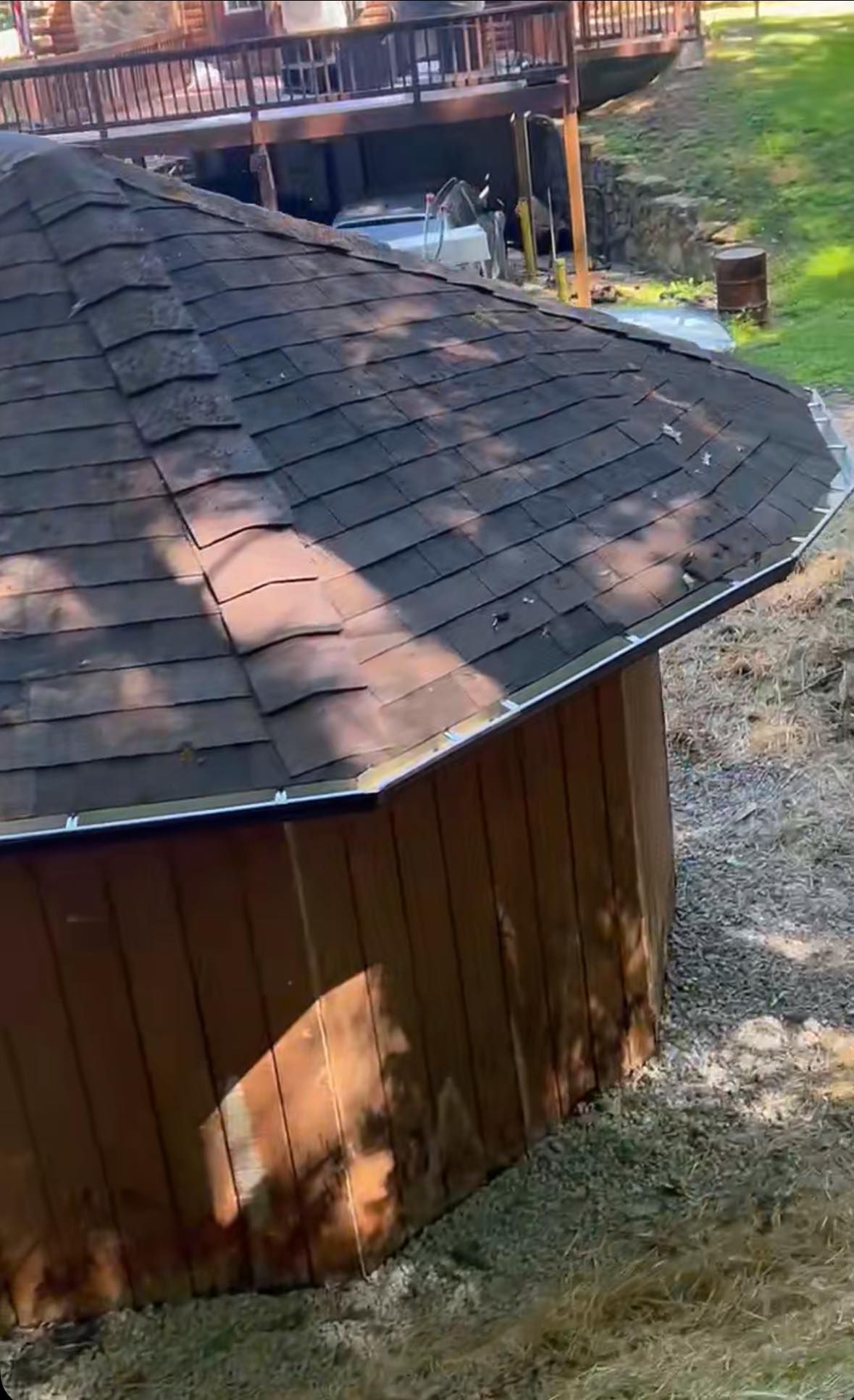A round structure with a shingled roof and vertical wooden siding, partially shaded, situated near a house and lawn.