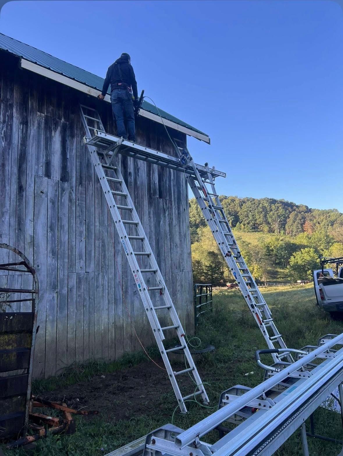 A person stands on a makeshift elevated platform supported by two ladders against the side of a wooden barn.