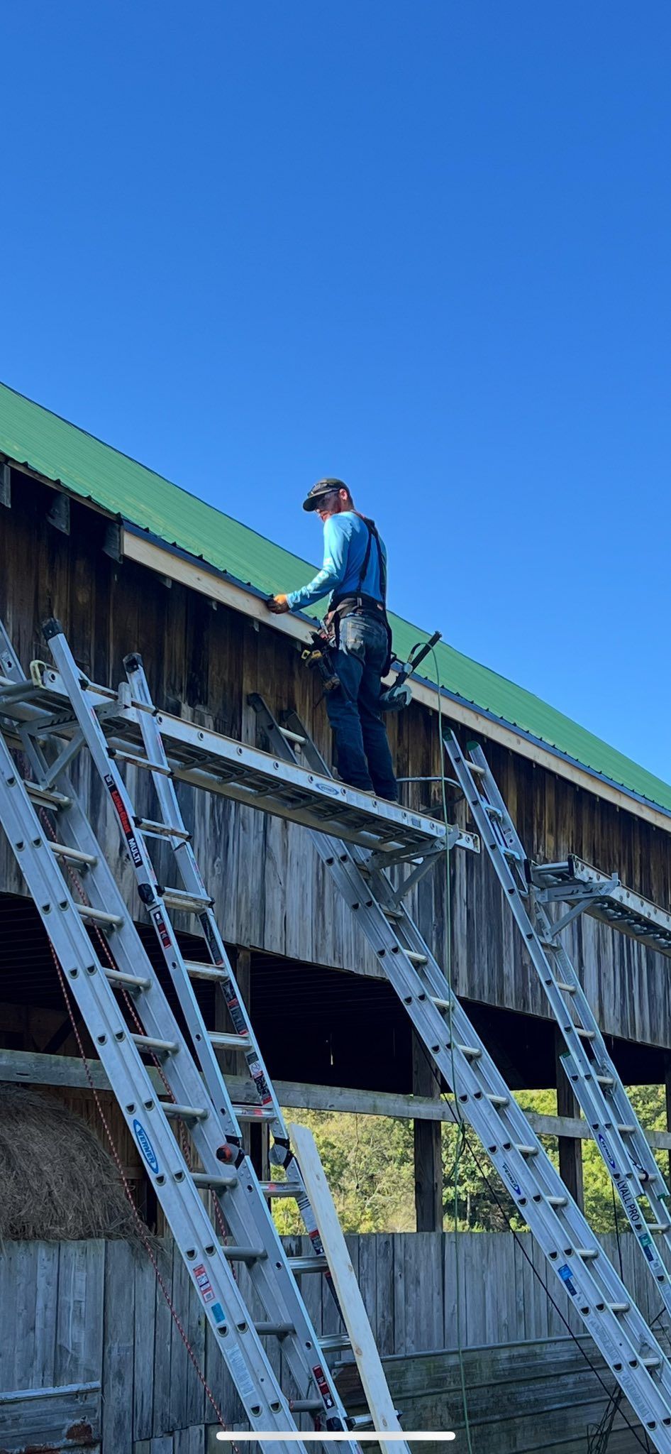 A person in a blue shirt works on the edge of a green barn roof while standing on a tall scaffolding platform.