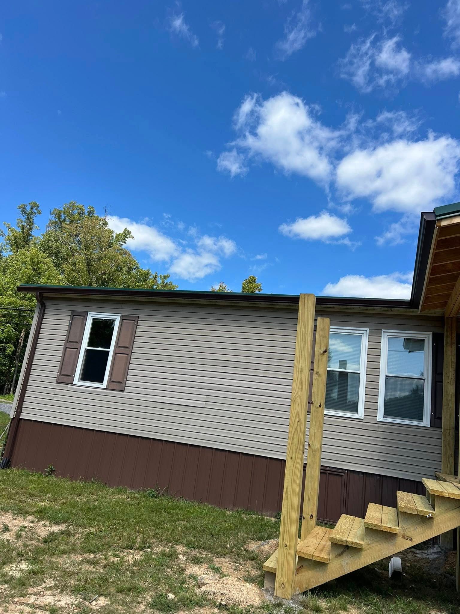 A mobile home with beige diamond-patterned siding and brown skirting under a bright blue sky with a wooden staircase.