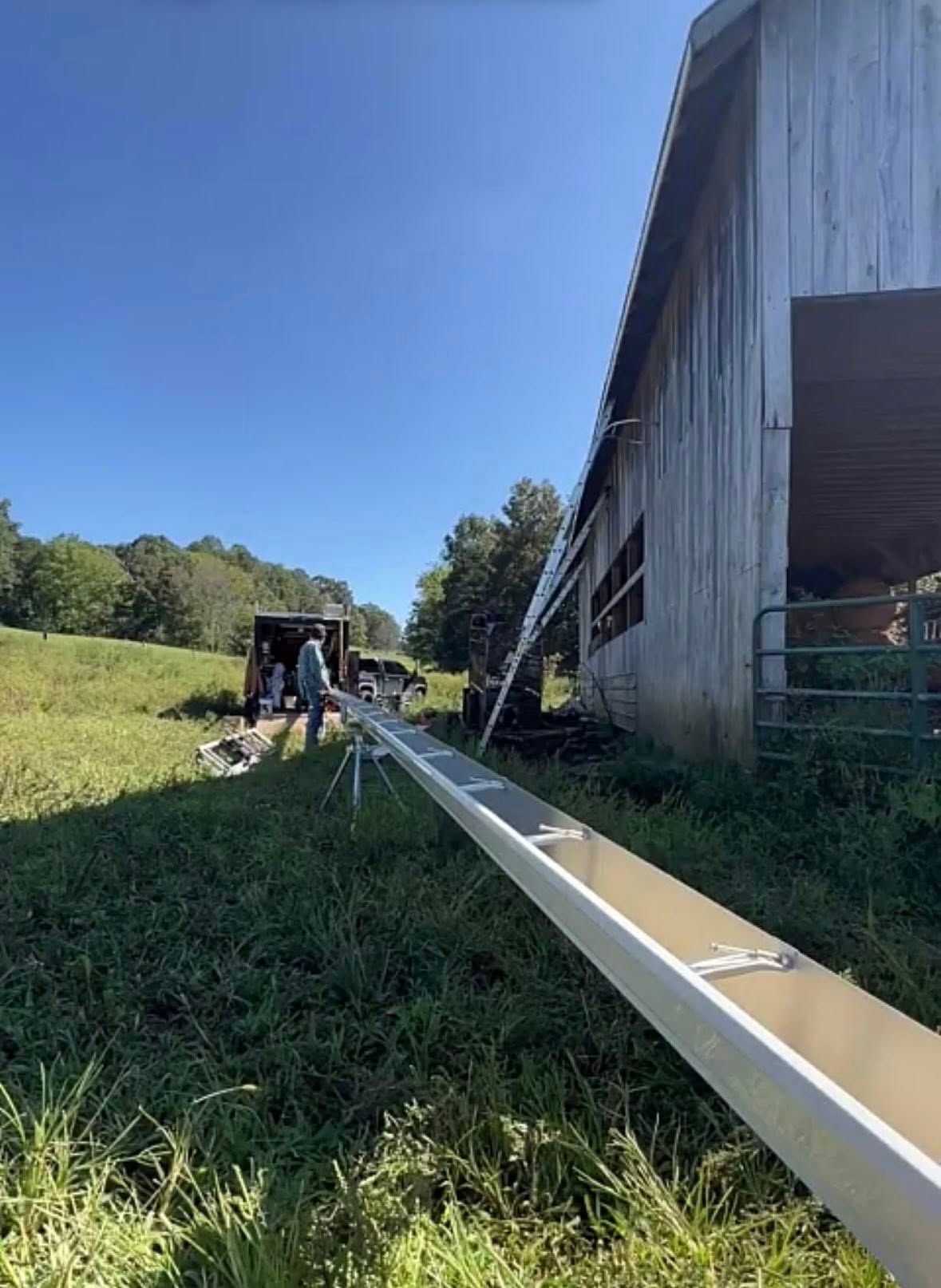 A long metal gutter section rests on a stand in a grassy field next to a rustic barn.
