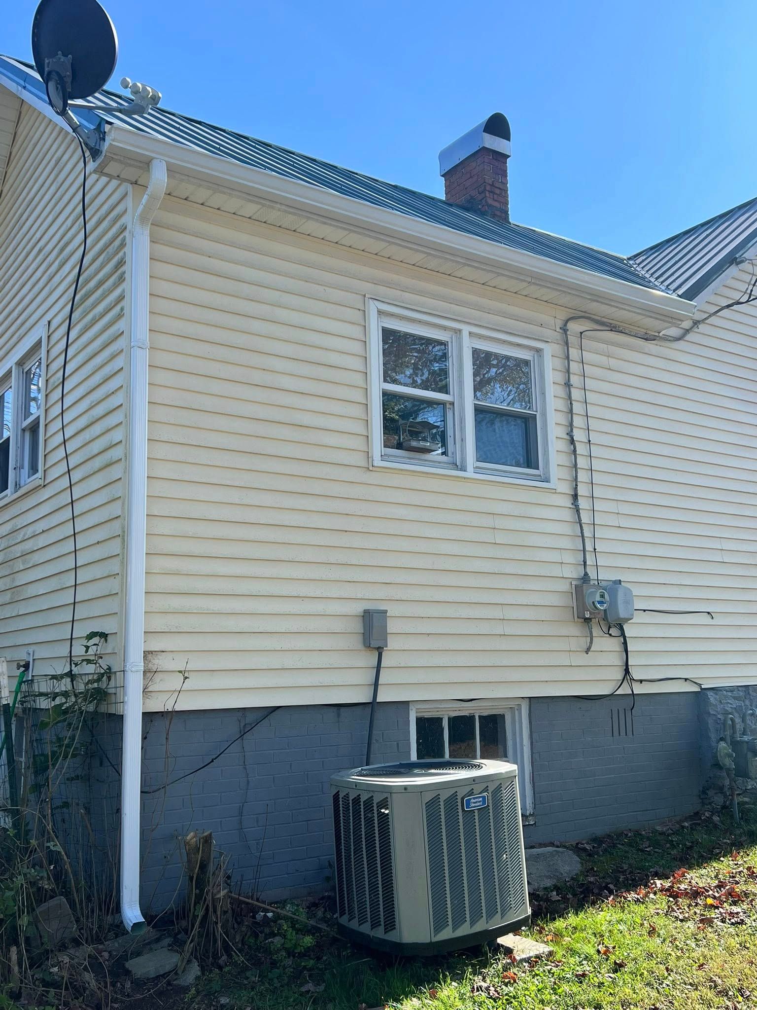 A pale yellow vinyl-sided house with a gray concrete foundation, an AC unit, and a brick chimney under a blue sky.