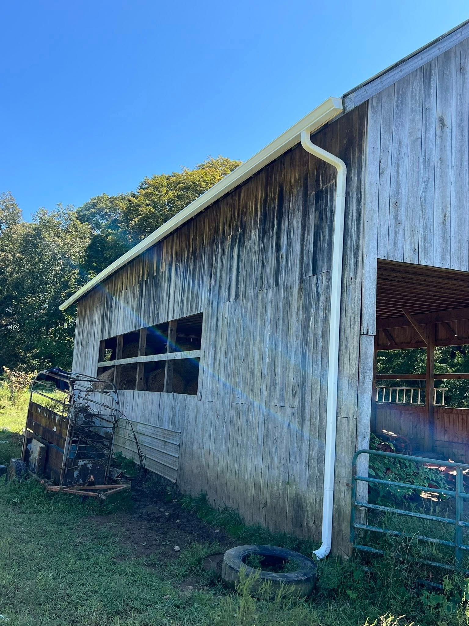 A side view of a weathered wooden barn with a newly installed white gutter and downspout, set against a clear blue sky.