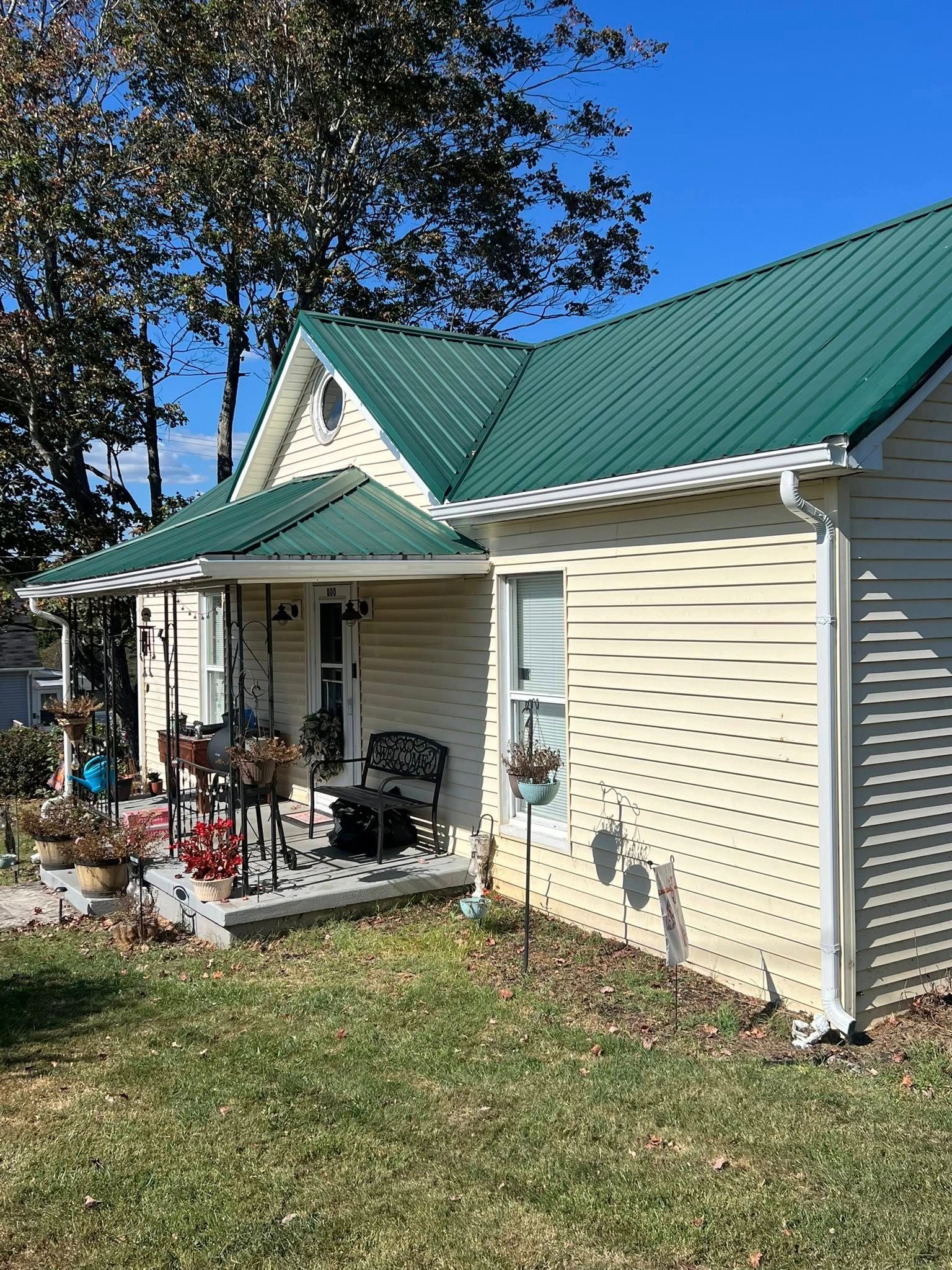 A yellow, single-story house with a green metal roof and a small front porch, surrounded by trees under a clear blue sky.