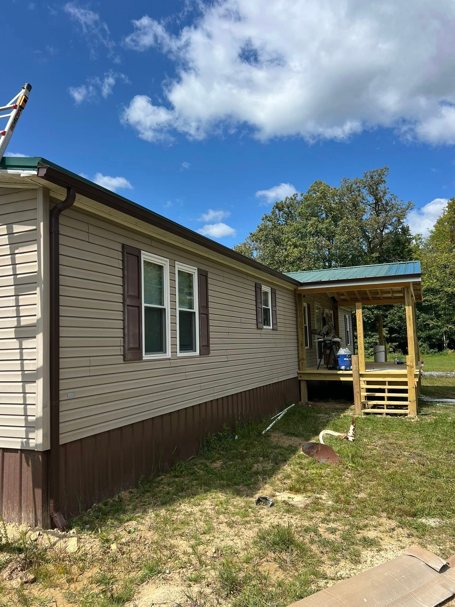 A side view of a tan mobile home with brown skirting, dark shutters, and a newly constructed wooden porch under blue sky.