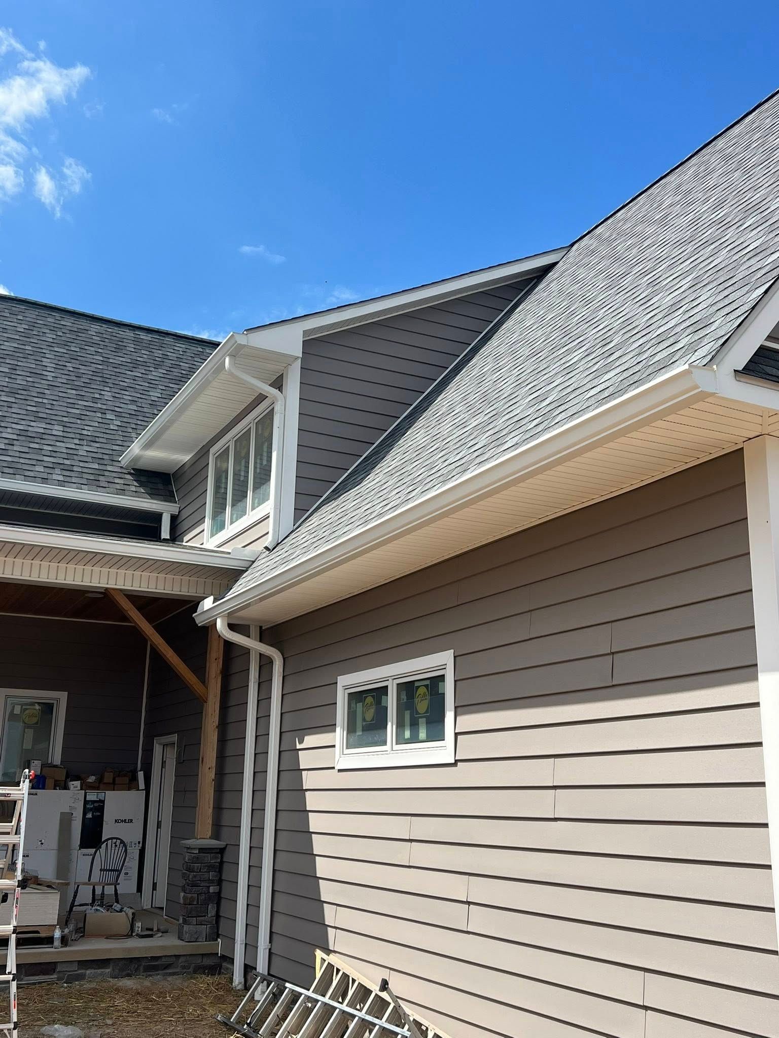 A side view of a house under construction, showing gray horizontal siding, a shingled roof, and a covered porch area.