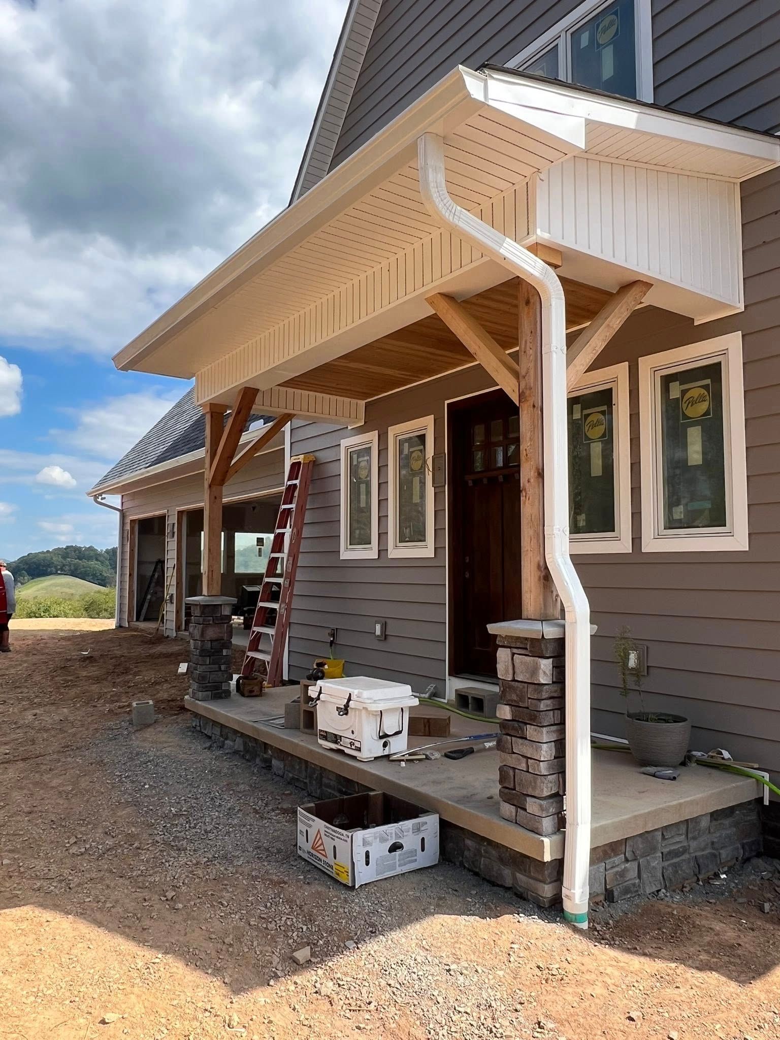 A front porch under construction, featuring wooden support posts on stone pillars, brown siding, and a white downspout.
