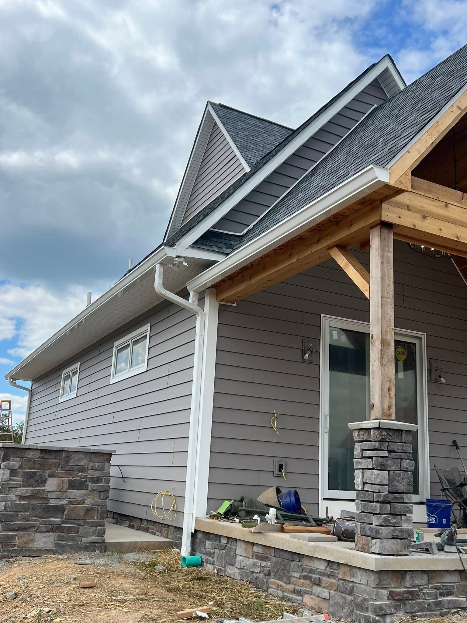 Exterior view of a house under construction with grey siding, stone accents, a wooden porch post, and a white downspout.