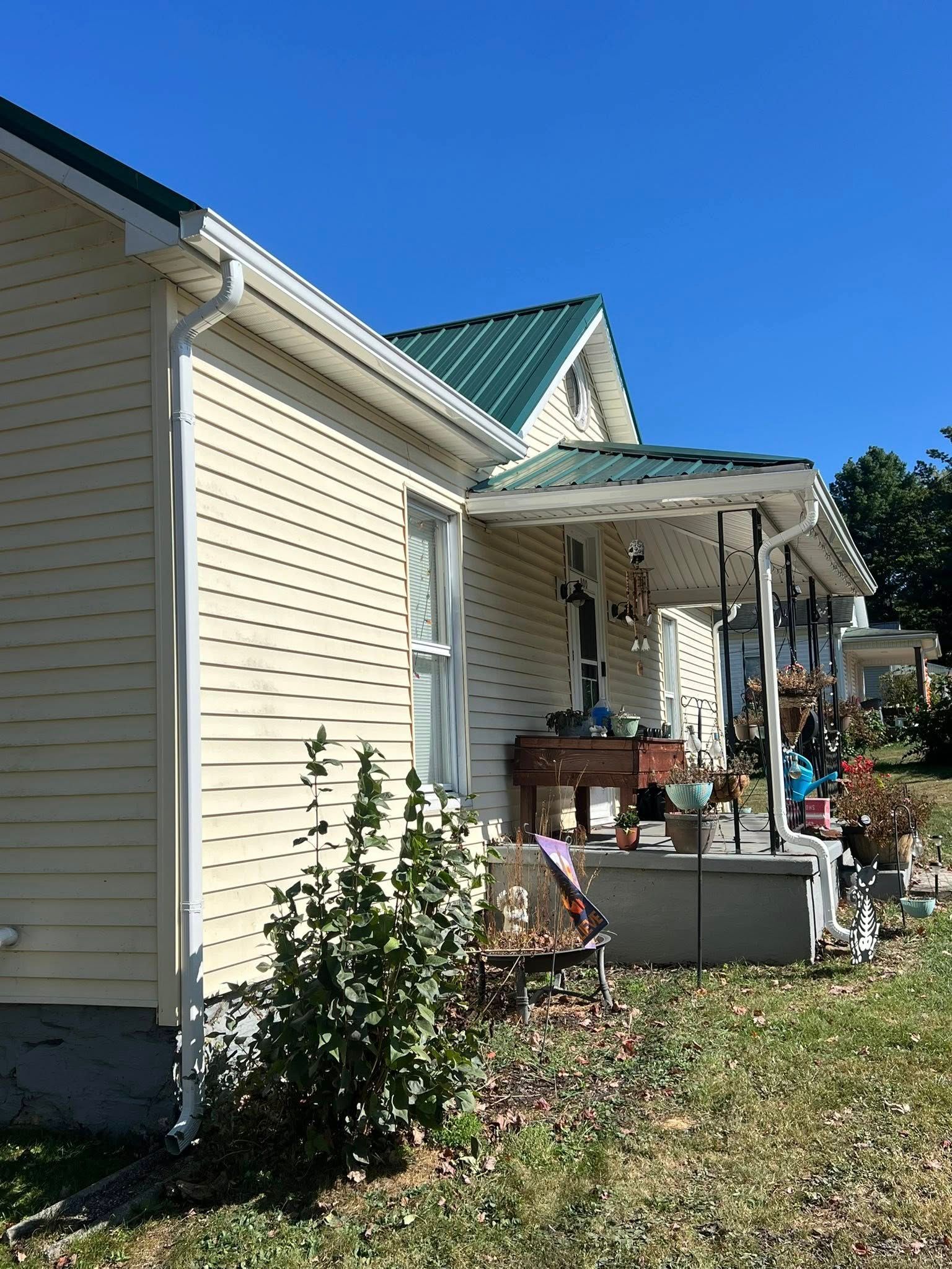 A light yellow house with a green metal roof, featuring a covered front porch decorated with various plants and items.