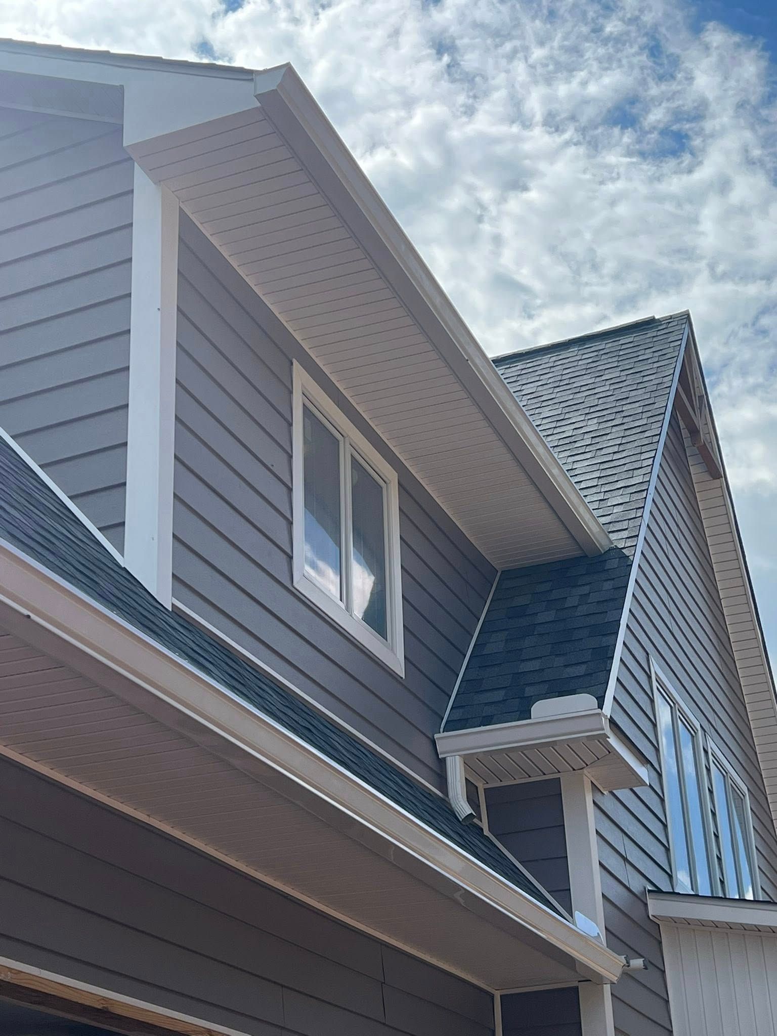 A low-angle view of a grey-sided house exterior with a white-trimmed window, dark shingled roofs, and a cloudy sky.