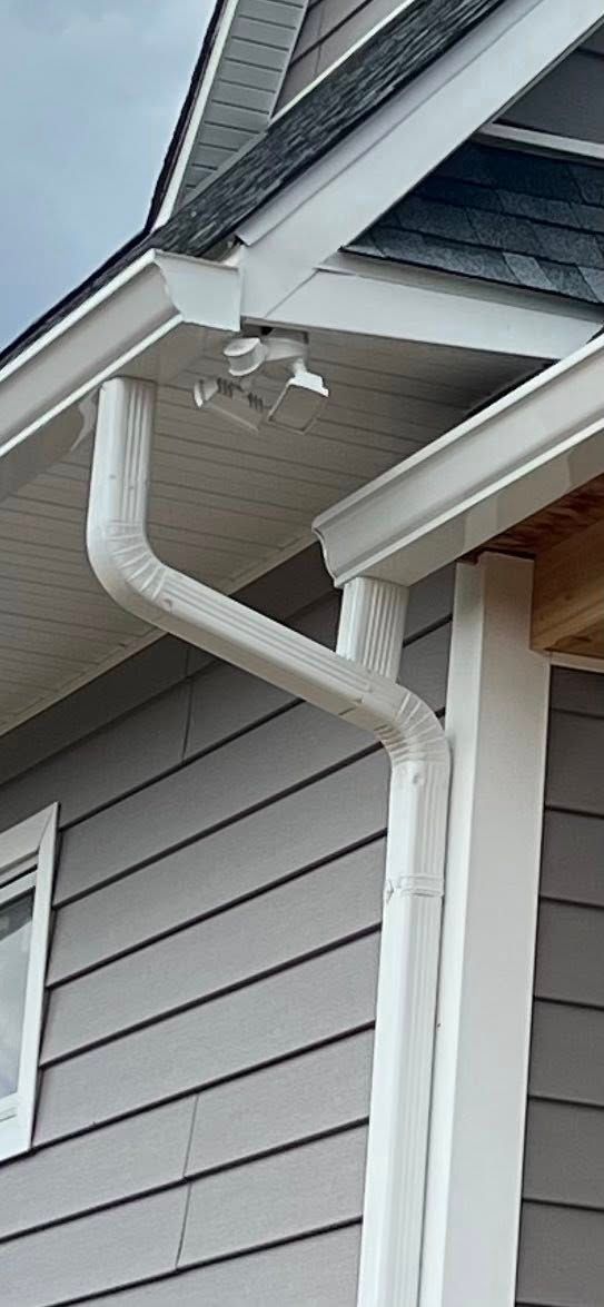 White gutter downspouts run along the corner of a grey-sided house, beneath an overhanging roof with a security light.