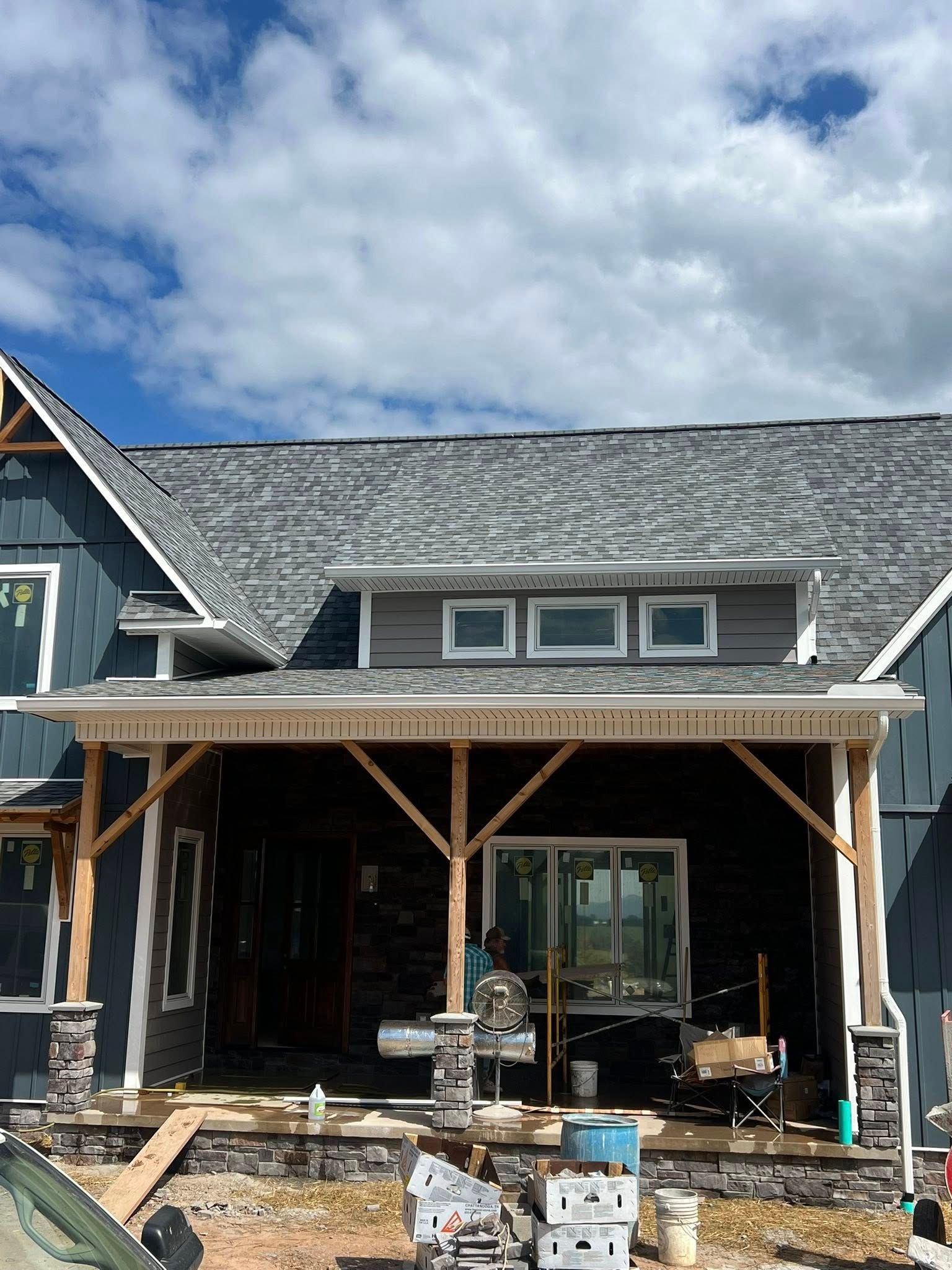 A house under construction with a front porch, grey shingles, blue siding, stone pillars, and a partially built entrance.