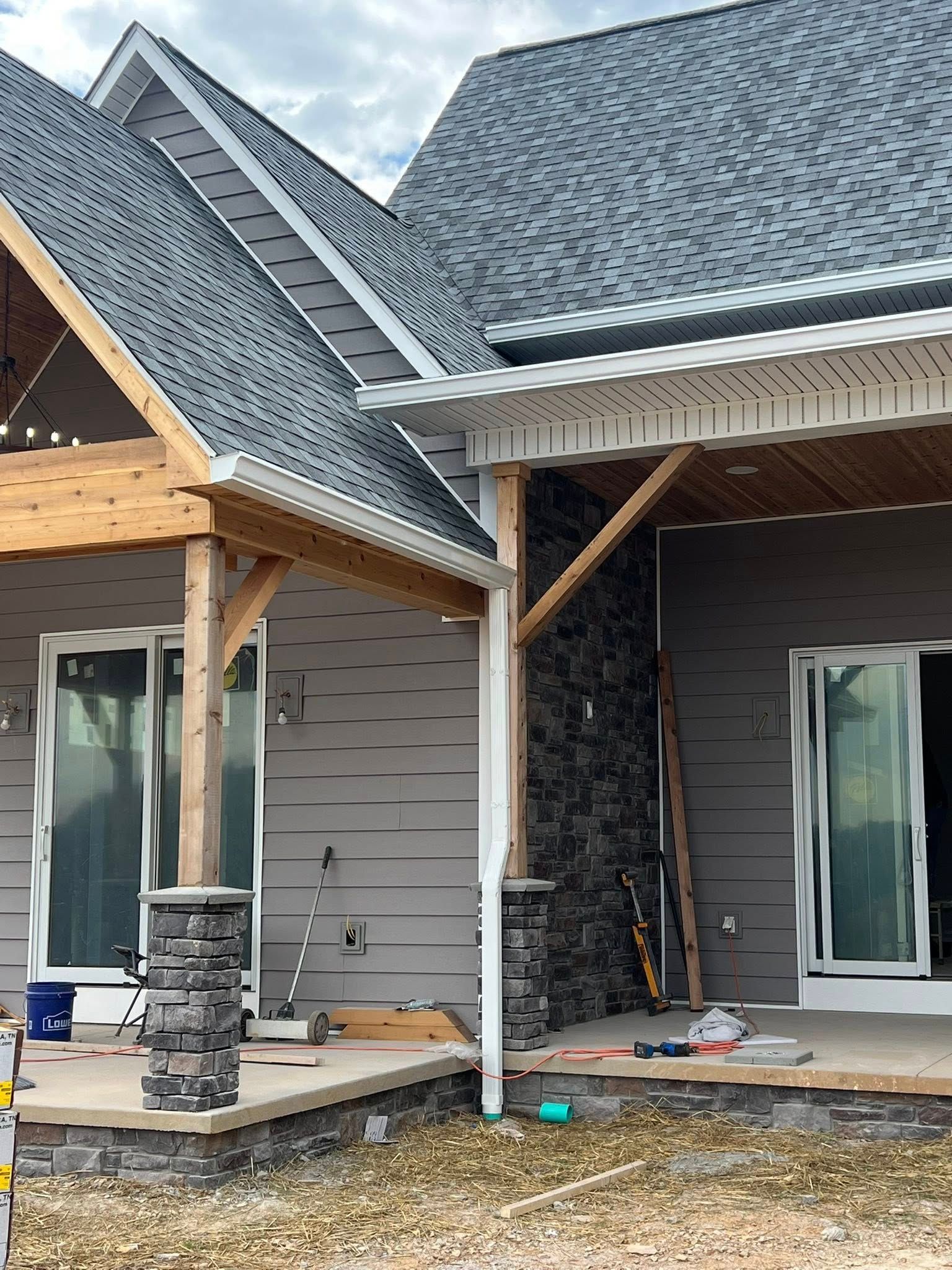 A partially constructed house with a gray roof, stone column bases, and wood framing, viewed from a gravel driveway.