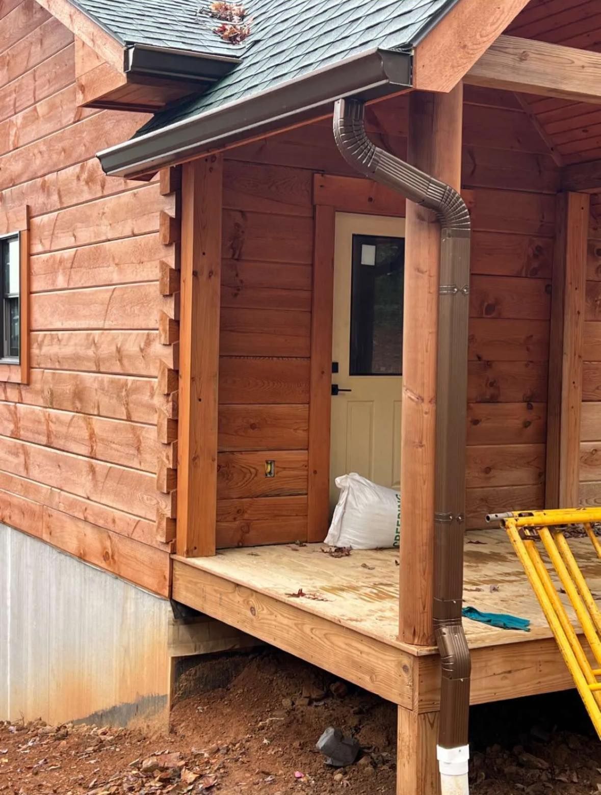 A brown log cabin entrance featuring a covered porch, a beige door, and a bronze gutter downspout on a wooden post.