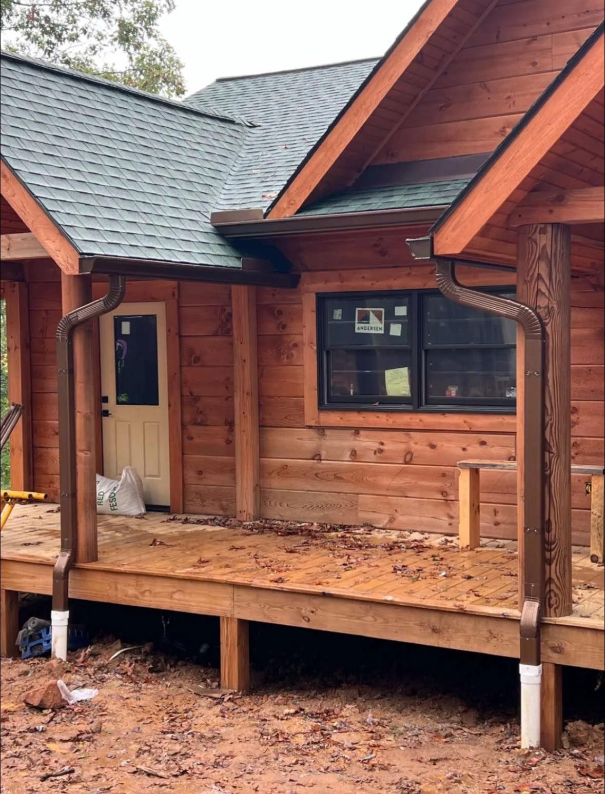 A rustic log cabin porch featuring dark wood siding, a green shingled roof, two brown downspouts, and a window with trim.