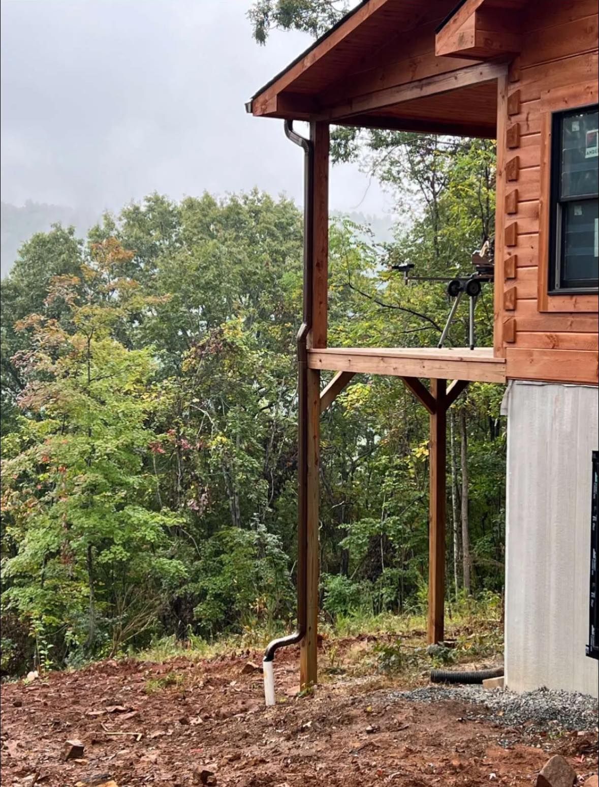 A side view of a wooden cabin on a hillside with a dark metal downspout against a backdrop of trees in fog.