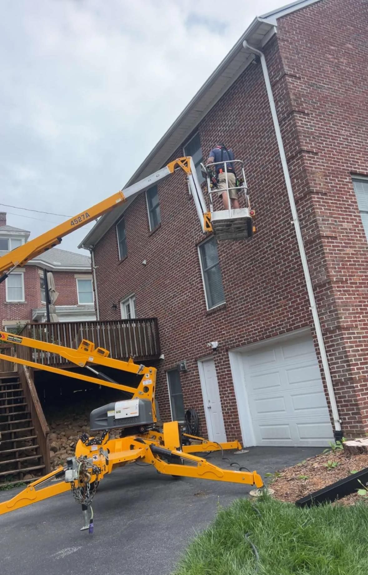 A person in a yellow aerial lift bucket works on the exterior siding of a two-story red brick house.