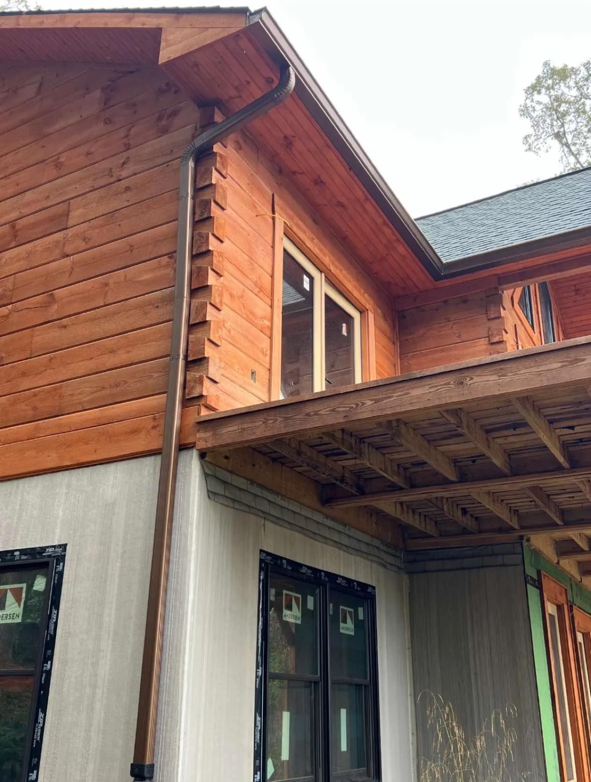 A low-angle view of a wooden log cabin under construction, featuring a copper downspout against a white foundation wall.