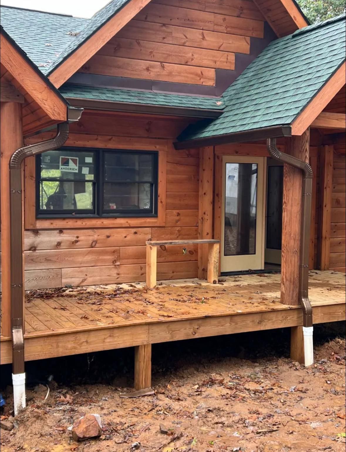 A wooden cabin porch with dark-framed windows, a glass door, and green roof shingles over a dirt-covered foundation.
