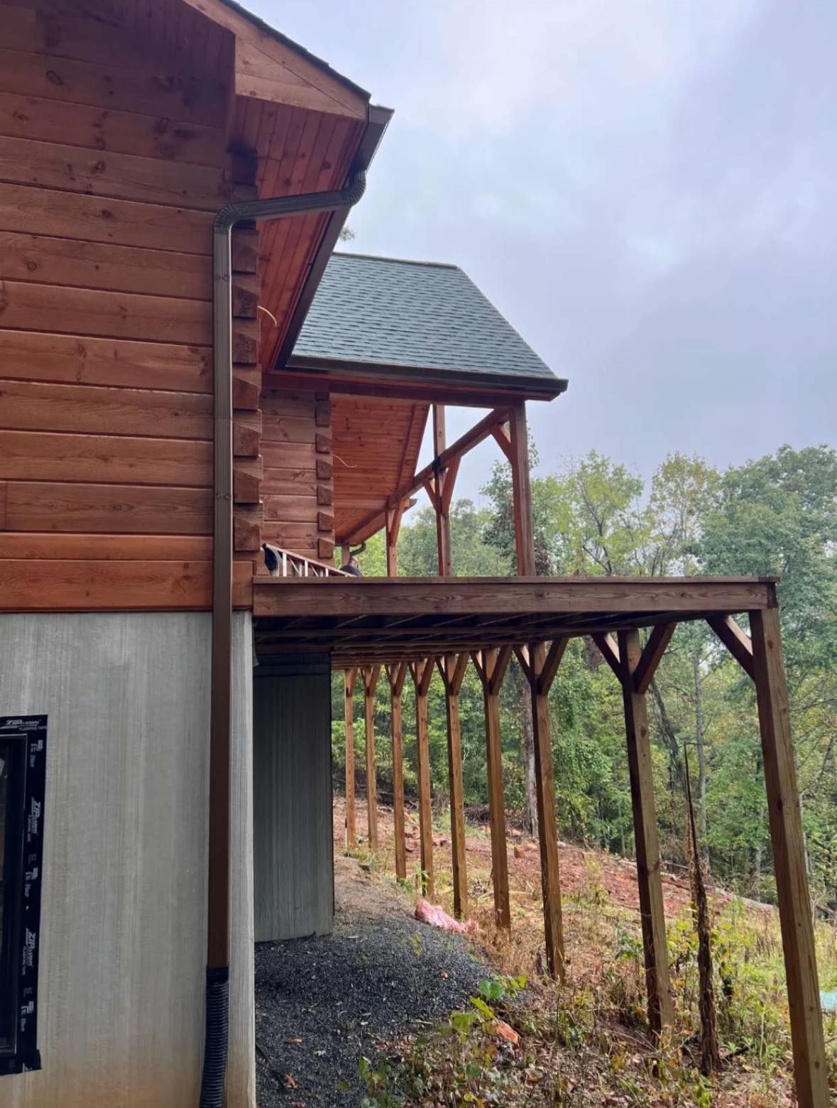 A side view of a brown log cabin with a concrete foundation, an elevated wooden deck, and a dark brown downspout.