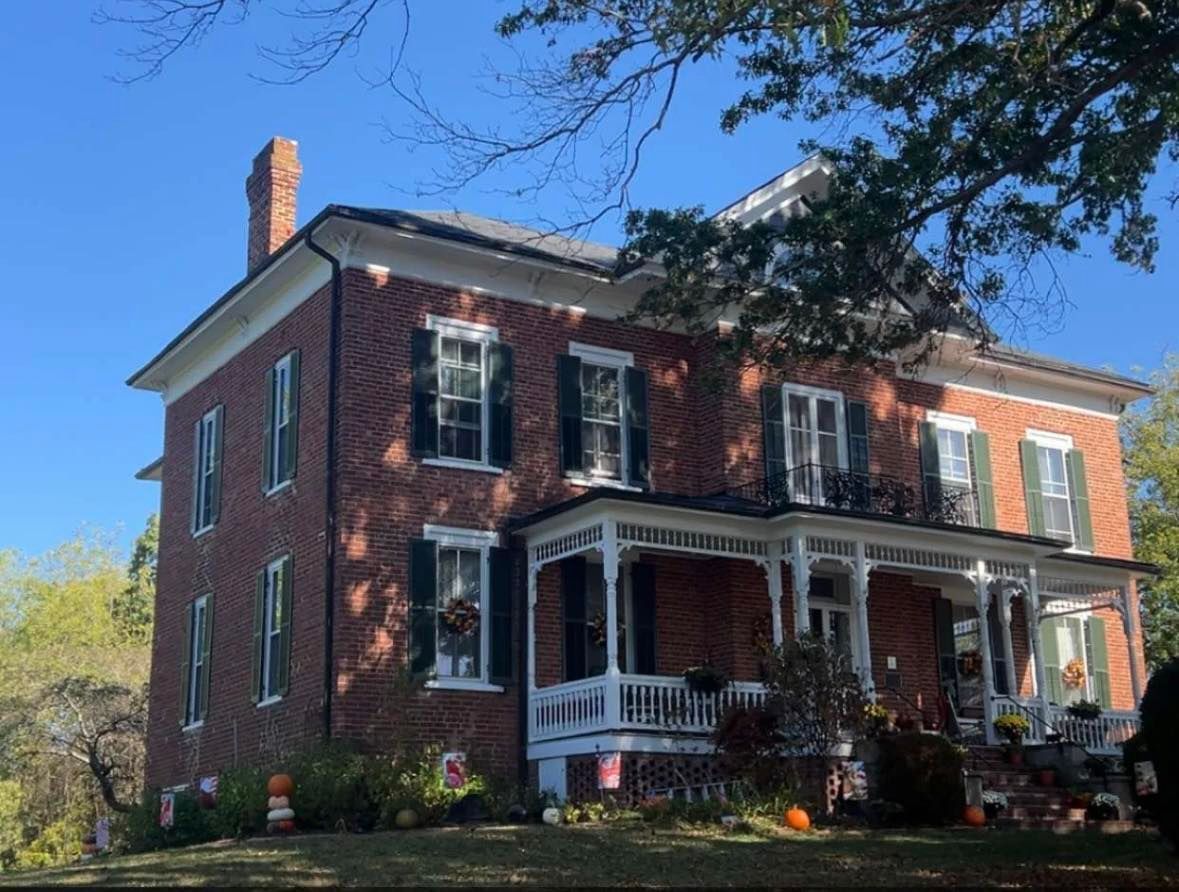 A two-story brick house with a white wrap-around porch, green shutters, and a chimney set under a clear blue sky.