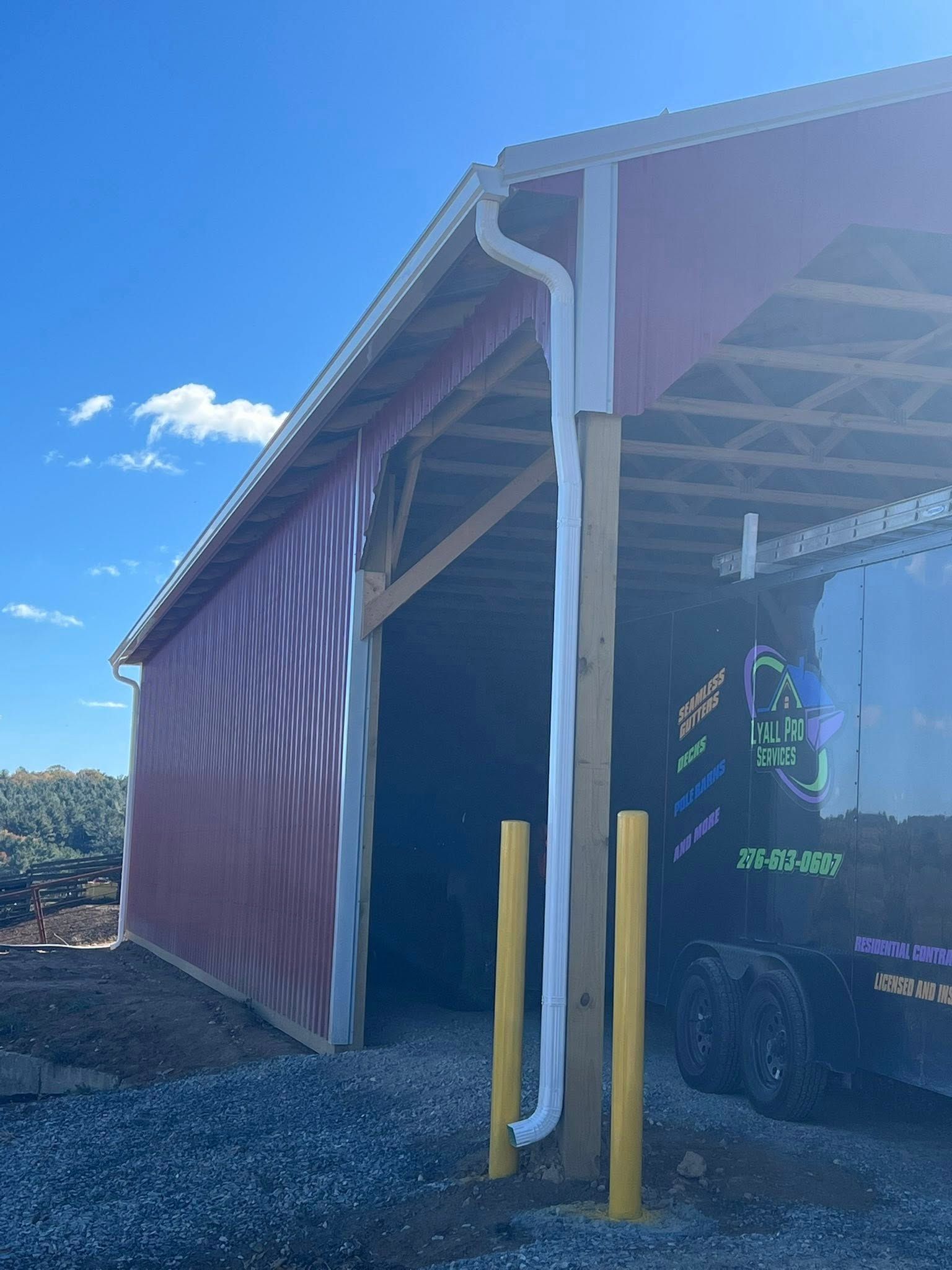 A white downspout runs down a wooden support beam on the corner of a red barn next to a parked black trailer.