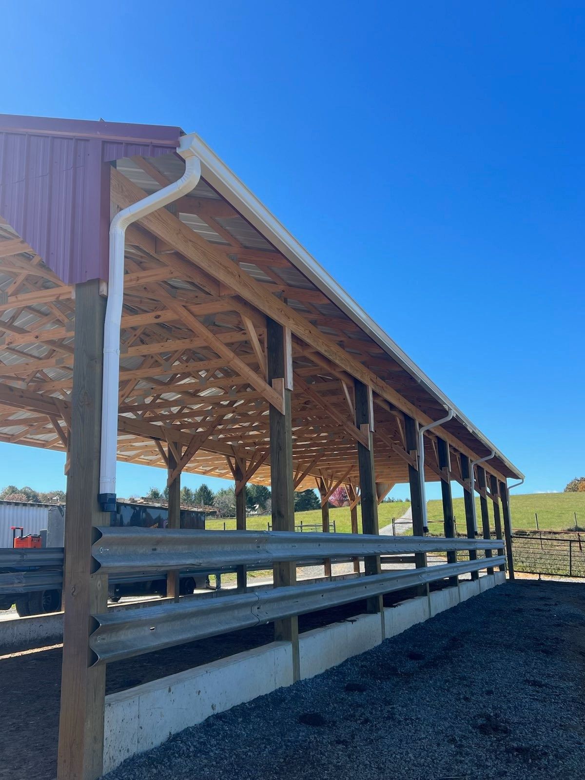An open-sided timber pole barn with a red roof, white gutters, and steel guardrails along the sides on a gravel base.