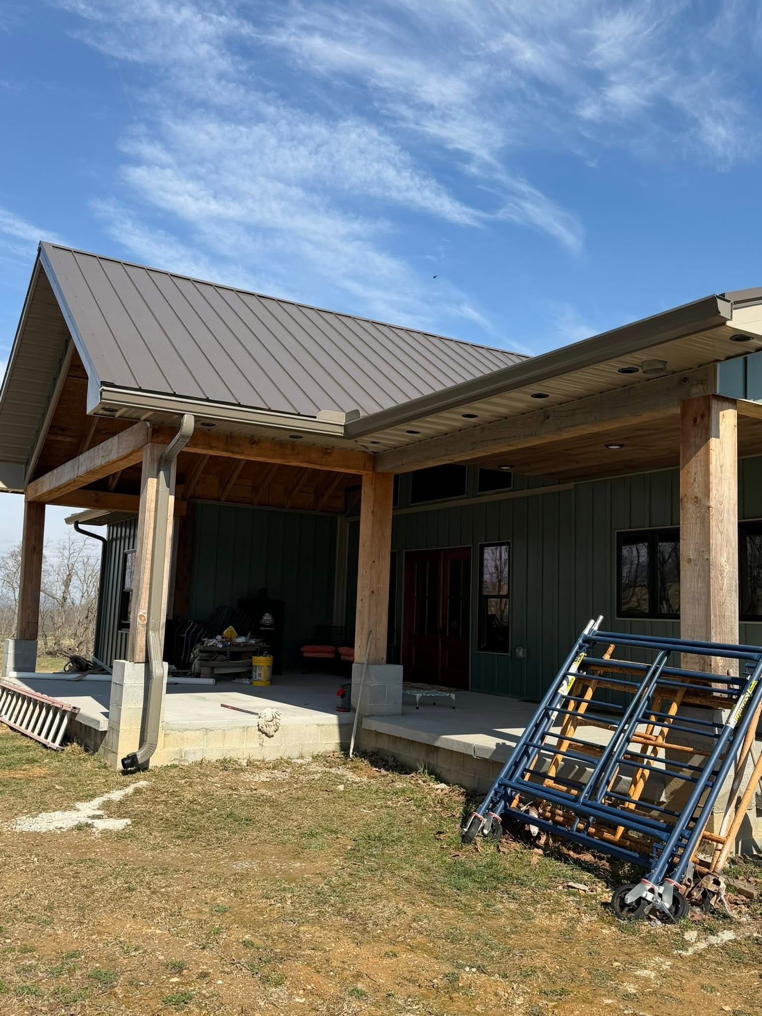 A modern home under construction with a metal roof, log-style support beams, and a concrete patio under a covered porch.