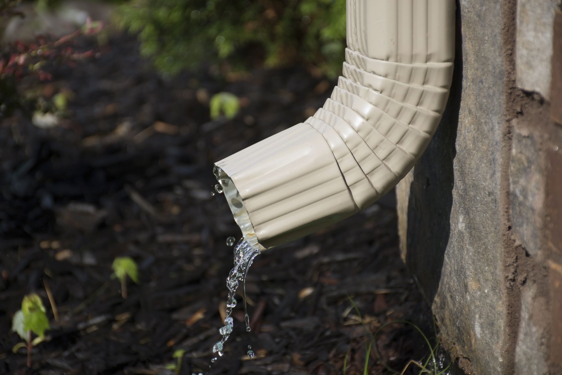 A tan metal downspout attached to a stone wall, draining water into a dark, mulched garden bed.