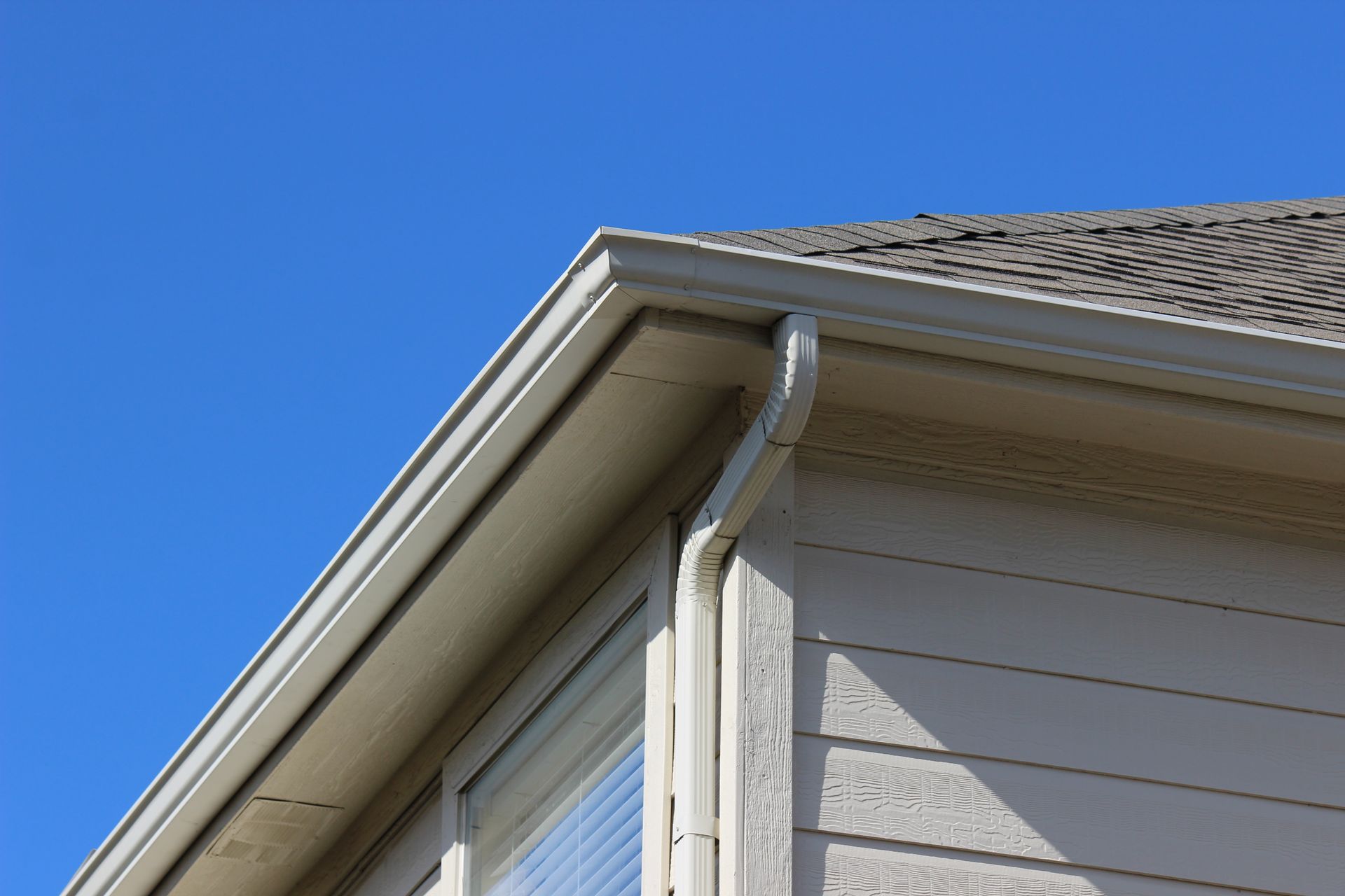 A low-angle view of a house roof edge with a gutter and downspout against a clear blue sky.