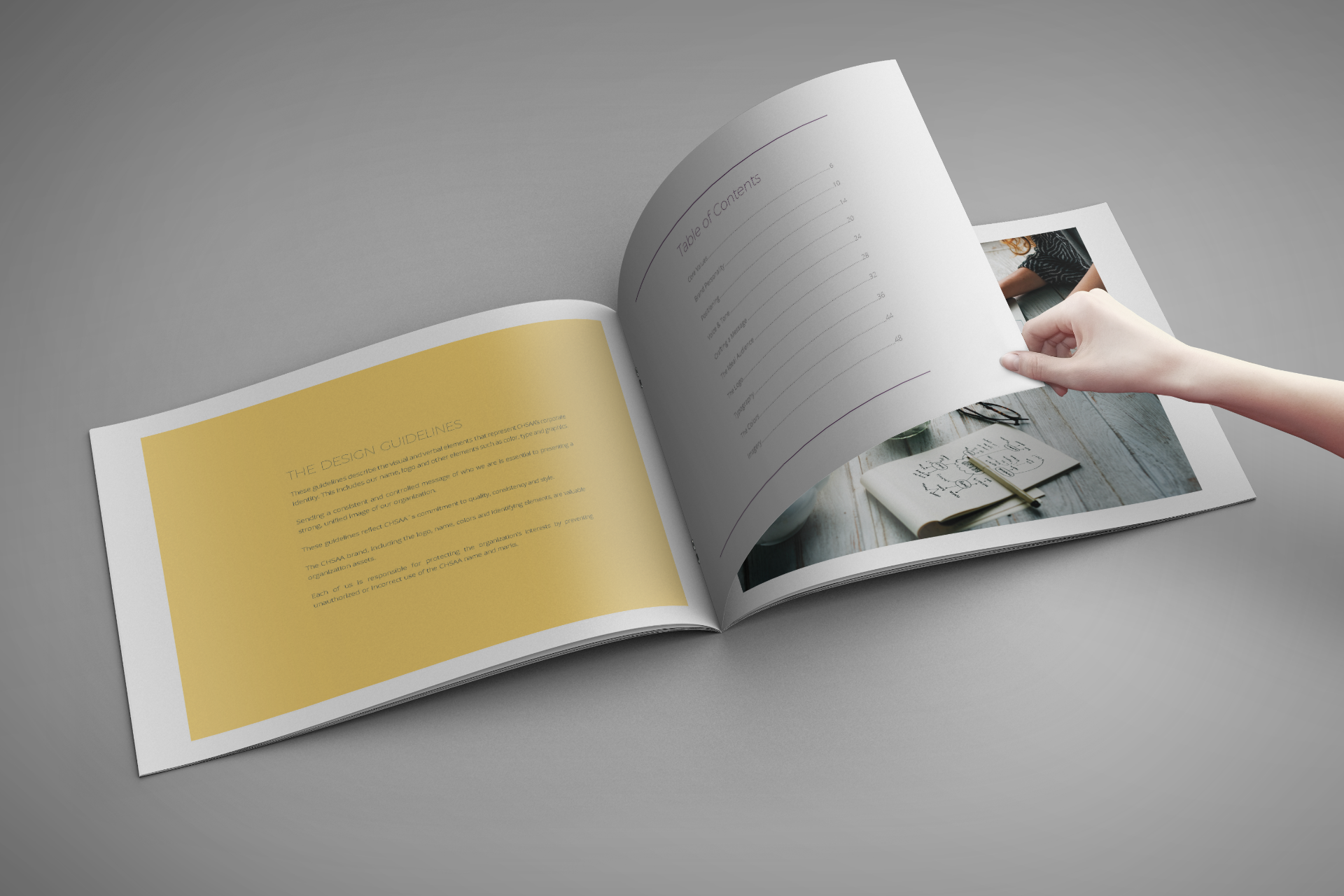 Open brochure with a hand turning the page, on a gray surface. Yellow and photo pages visible.