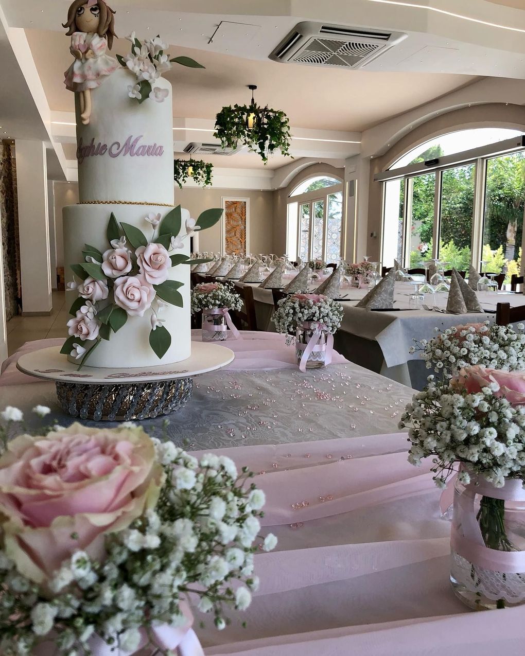 Una torta alta a due piani, bianca, sormontata da una bambola e decorata con rose rosa, posta su un tavolo con centrotavola floreali.