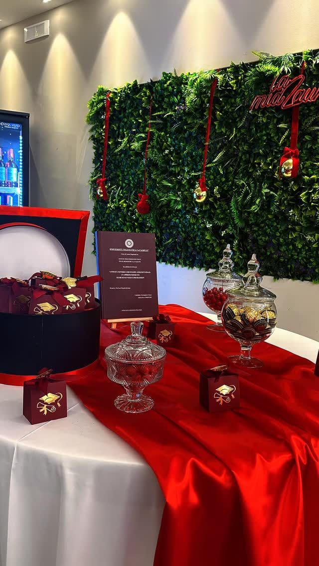 Una tavola apparecchiata per un evento formale con un runner rosso, barattoli di vetro contenenti dolcetti, scatole regalo e uno sfondo decorativo verde.