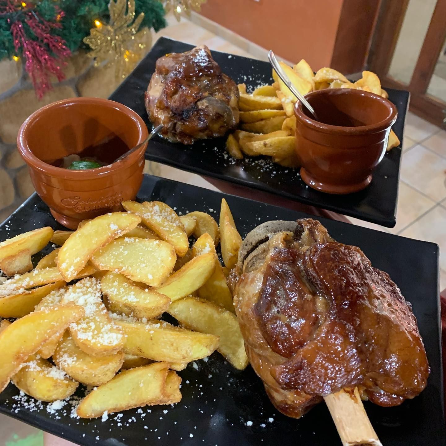 Due piatti rettangolari neri contengono stinchi di maiale arrostiti serviti con spicchi di patate speziate e salsa per intingere.