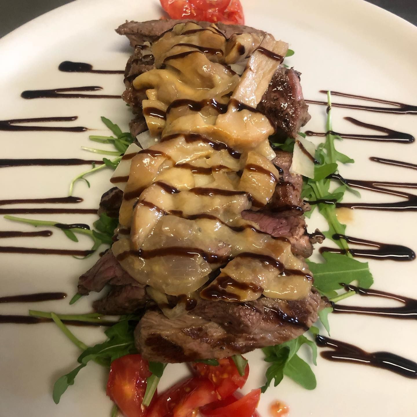 Fettine di bistecca guarnite con funghi e glassa balsamica, servite su un letto di rucola con spicchi di pomodoro su un piatto bianco.