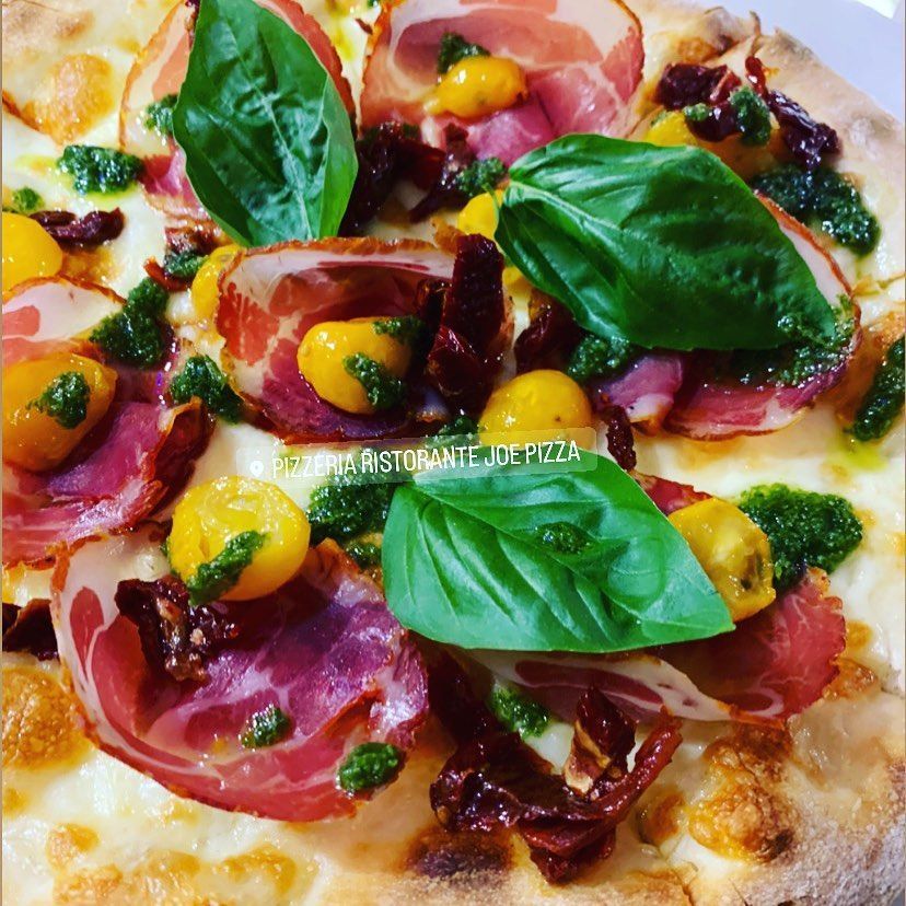 Pizza gourmet condita con salumi, pomodori gialli, foglie di basilico fresco e pesto verde su una base sottile e dorata.