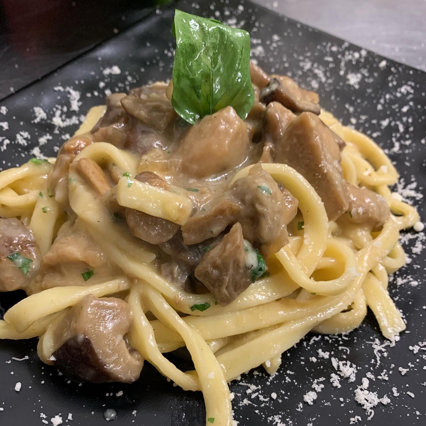 Una ciotola di pasta ai funghi in salsa cremosa, guarnita con basilico fresco e parmigiano, servita su un piatto nero.