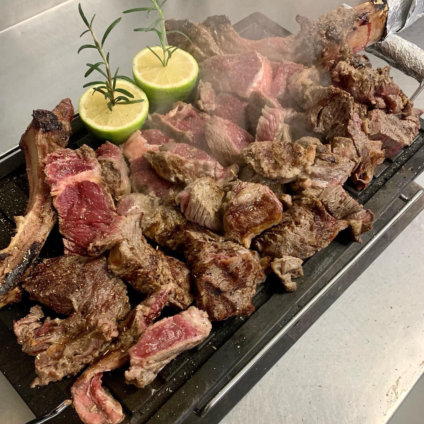 Un piatto di bistecca grigliata a fette, servito su una superficie metallica con due mezze limoni e rametti di rosmarino fresco.